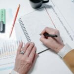 Dłoń robiąca notatki w zeszycie leżącym obok wykresów na biurku