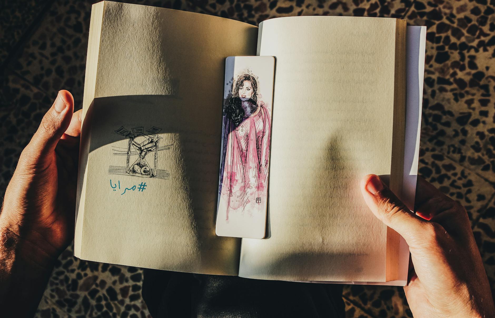 Dłonie trzymają otwartą książkę z artystyczną zakładką w słońcu