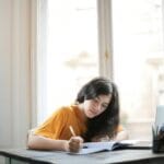 Młoda kobieta pisze notatki przy oknie, przygotowując rozprawkę z literatury