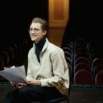 Aktor uśmiecha się, siedząc na scenie pustego teatru z tekstem sztuki
